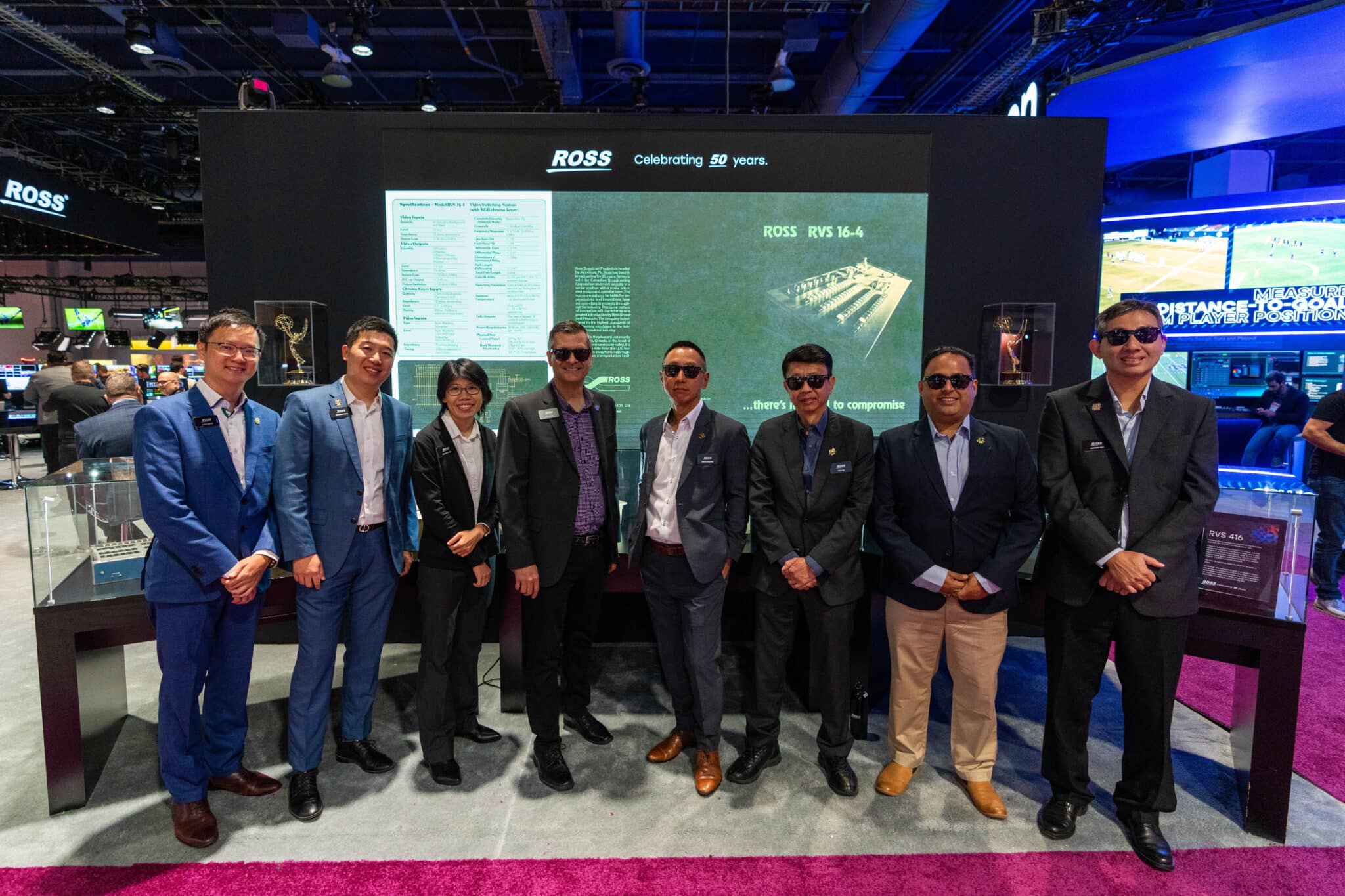 Group of people in suits wearing sunglasses stnadingin in front of large TV screen that says Ross, celebrating 50 years at a convention